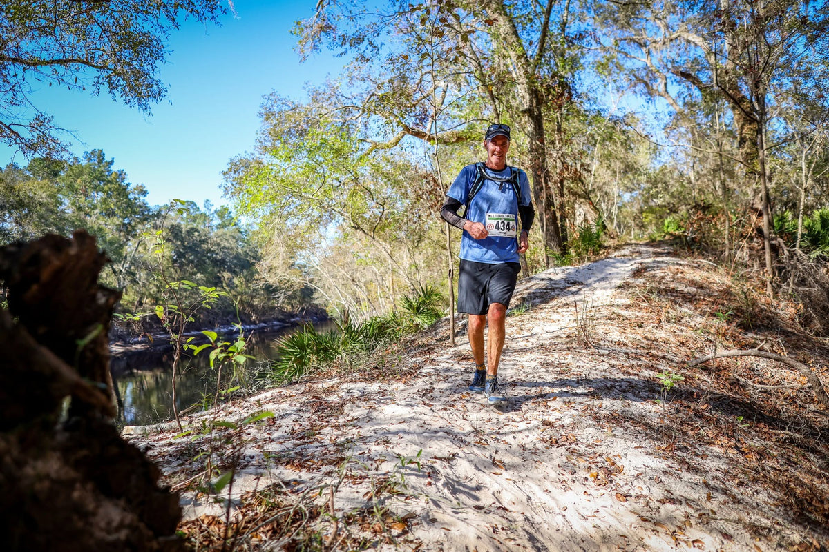 A male trail runner wearing a blue long-sleeve shirt, black shorts, and a hydration pack runs along a sandy path surrounded by trees and a river under a blue sky.