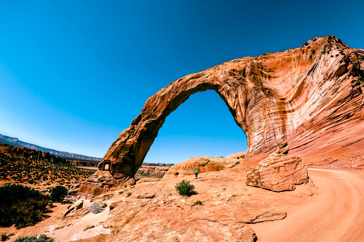 A scenic landscape photograph of a large natural sandstone arch under a clear blue sky, with red rock desert terrain and a dirt road in the foreground.