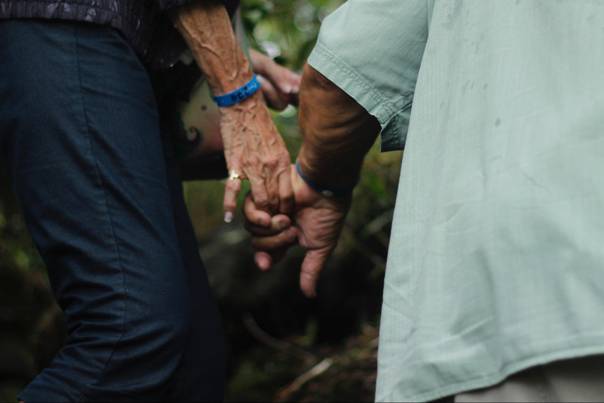 Two people holding hands in an outdoor wooded setting. One person wears a dark patterned top and dark pants, and the other wears a light mint green shirt.