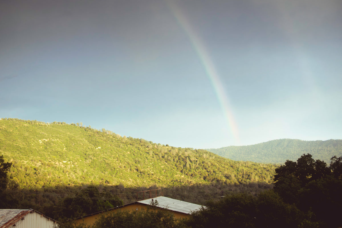 A scenic landscape photograph of green forested mountains under a blue sky with a faint rainbow arching across the upper center.